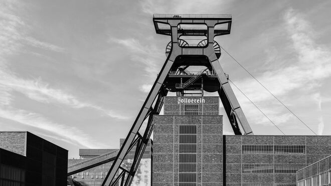 Das Bild zeigt das markante Fördergerüst der ehemaligen Zeche Zollverein in Essen. Die Schwarz-Weiß-Aufnahme betont die klaren Linien und die industrielle Architektur des Stahlbaus. Im Vordergrund dominiert das große, geometrische Fördergerüst, während di