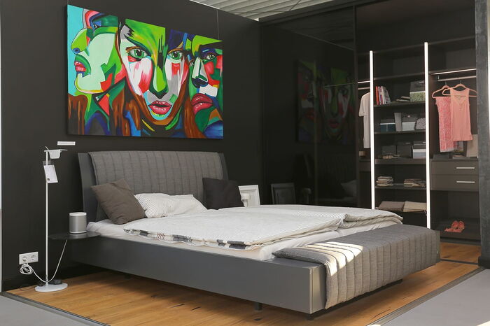 Ein modernes Schlafzimmer  mit einem grauen Bett. An der Wand hängt ein großes expressionistisches Bild mit Gesichtern in bunten Farben.