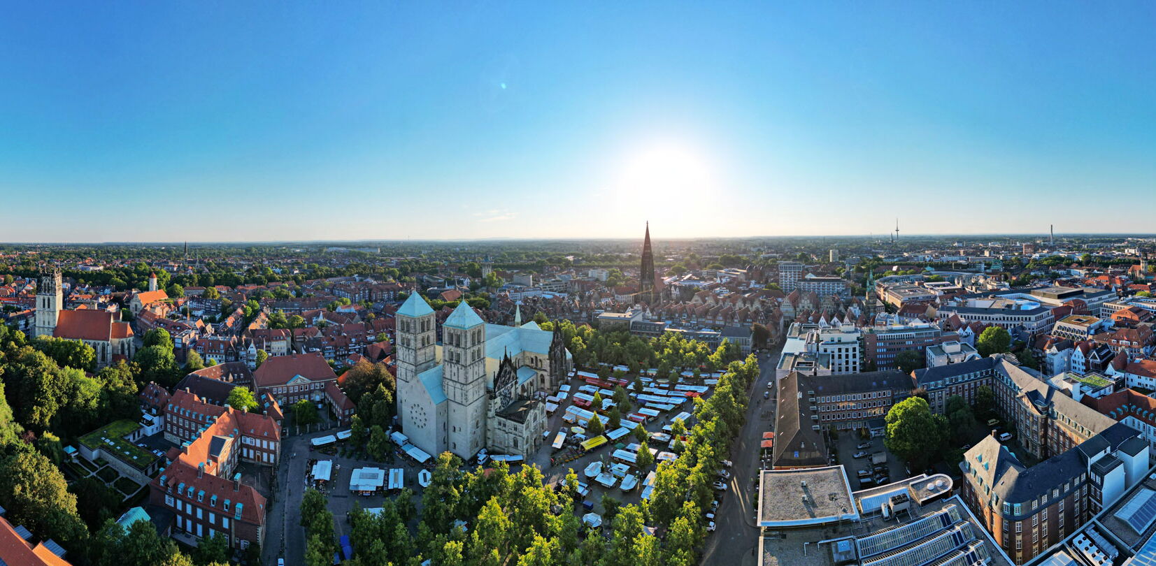 Das Panorama zeigt die Stadt Münster aus der Vogelperspektive bei strahlendem Sonnenschein. Im Zentrum des Bildes fällt der St.-Paulus-Dom mit seinen markanten Türmen auf. Rundherum erstreckt sich die Altstadt mit ihren roten Ziegeldächern, historischen G