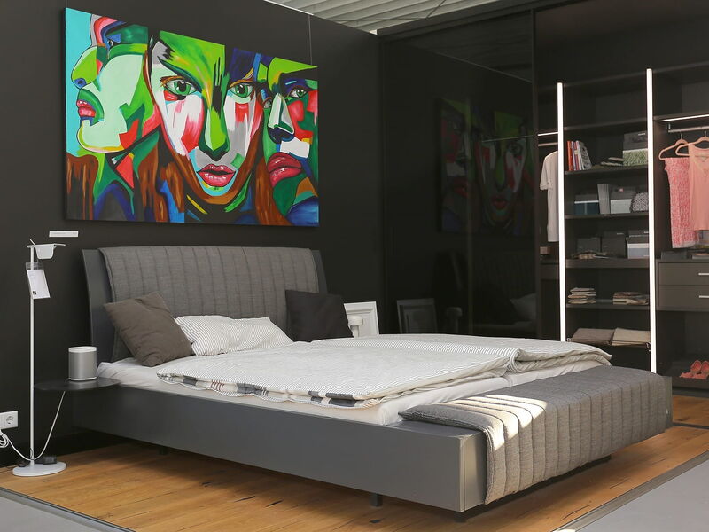 Ein modernes Schlafzimmer  mit einem grauen Bett. An der Wand hängt ein großes expressionistisches Bild mit Gesichtern in bunten Farben.