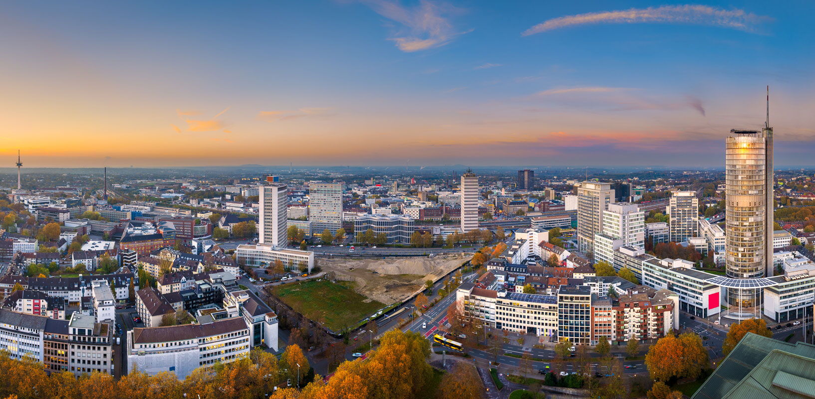 Das Panorama zeigt die Skyline der Stadt Essen bei Sonnenuntergang. Zu sehen sind zahlreiche Hochhäuser, Wohn- und Geschäftsgebäude sowie weitläufige Stadtbereiche, die sich bis zum Horizont erstrecken. Rechts im Bild steht ein markantes rundes Hochhaus,