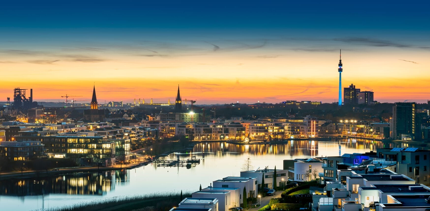Ansicht der Skyline von Dortmund bei Dämmerung mit Wasserfläche, Gebäuden und Fernsehturm im Hintergrund.